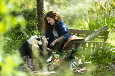 Frau sitzt auf Holzbank mit Hund im Garten - SBOF03808