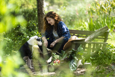 Woman sitting on wooden bench with dog in garden - SBOF03808