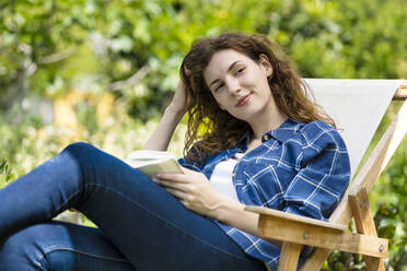 Beautiful woman with hand in hair holding book while relaxing on chair in garden - SBOF03810