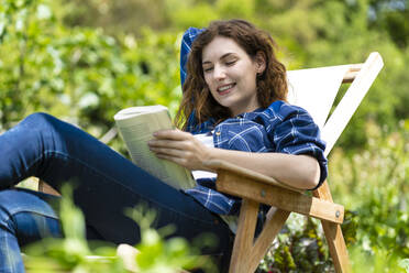 Lächelnde Frau liest ein Buch und sitzt auf einem Stuhl im Garten - SBOF03811