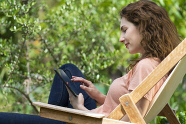 Young woman using digital tablet while sitting on chair in garden - SBOF03816
