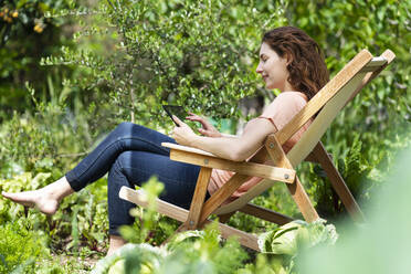 Frau sitzt auf einem Stuhl und benutzt ein digitales Tablet im Garten - SBOF03817