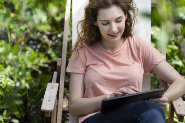 Junge Frau benutzt digitales Tablet auf einem Stuhl im Garten - SBOF03818