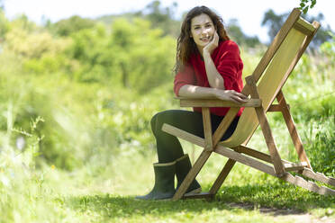 Schöne Frau mit Hand am Kinn sitzt auf einem Stuhl im Garten - SBOF03835
