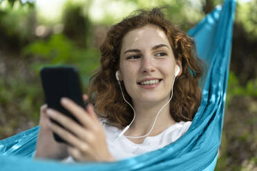 Smiling woman looking away listening music while relaxing on hammock - SBOF03847