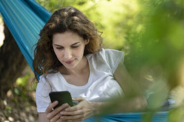 Junge Frau benutzt ihr Smartphone, während sie sich in der Hängematte entspannt - SBOF03848