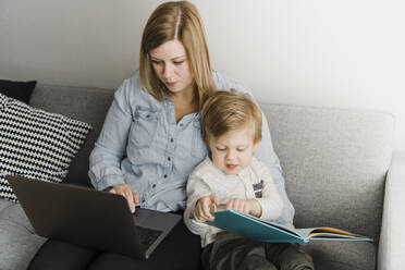 Mother using laptop while sitting son reading picture book on sofa at home - HWHF00027