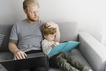 Der Sohn liest ein Bilderbuch, während der Vater mit dem Laptop zu Hause auf dem Sofa sitzt - HWHF00028