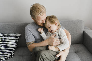 Playful father tickling son on sofa in living room - HWHF00029