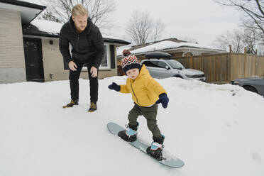 Mann schaut seinem Sohn zu, der vor dem Haus Snowboarden lernt - HWHF00034