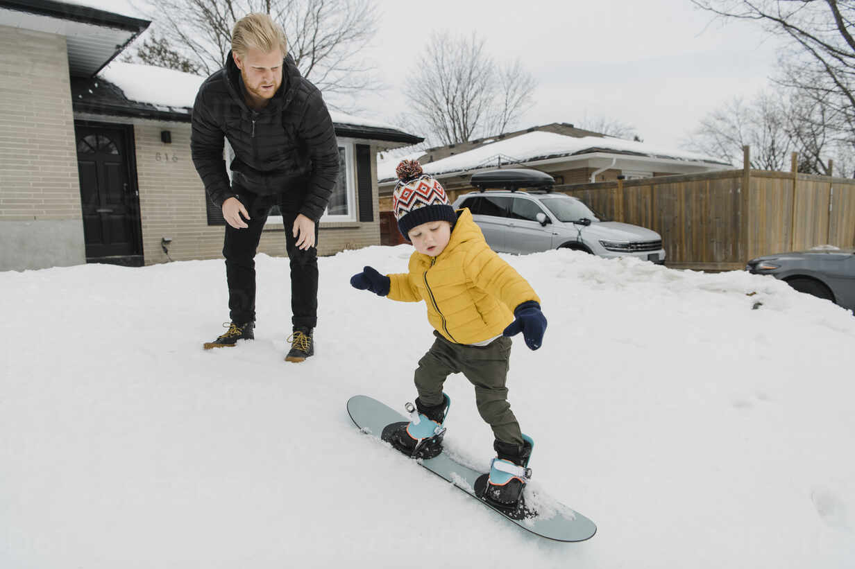 Man looking at son learning snowboarding outside house stock photo