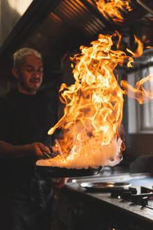 Chef with flame on frying pan in kitchen - JAQF00654