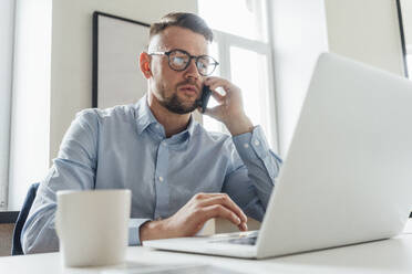 Mittlerer erwachsener Geschäftsmann mit Laptop, der im Büro mit einem Mobiltelefon spricht - VPIF04061