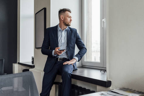 Männlicher Unternehmer, der ein Mobiltelefon hält, während er im Büro am Fenster sitzt - VPIF04085