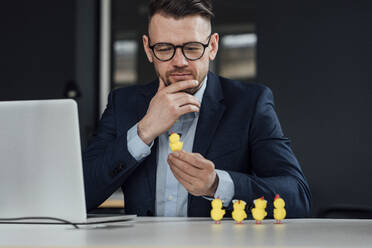 Nachdenklicher Geschäftsmann mit Hand am Kinn, der ein Baby-Huhn-Spielzeug im Büro betrachtet - VPIF04106