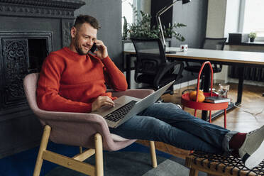 Männlicher Unternehmer, der einen Laptop benutzt, während er im Büro mit seinem Handy telefoniert - VPIF04111