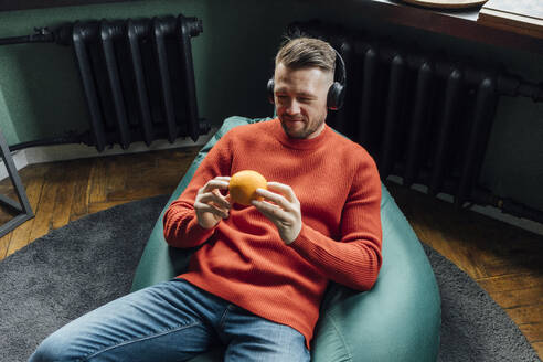 Lächelnder männlicher Unternehmer mit Kopfhörern, der eine orangefarbene Frucht betrachtet, während er auf einem Sitzsack sitzt - VPIF04118