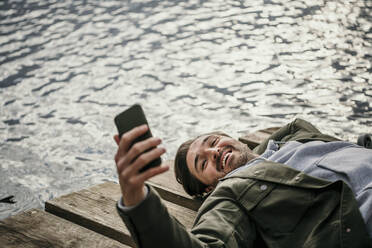 Smiling man using mobile phone while lying on jetty - MJRF00598