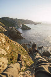 Personal perspective of man sitting at edge of coastal cliff at sunset - RSGF00694