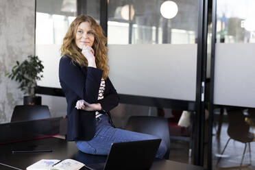 Businesswoman with hand on chin sitting on desk at office - FKF04160