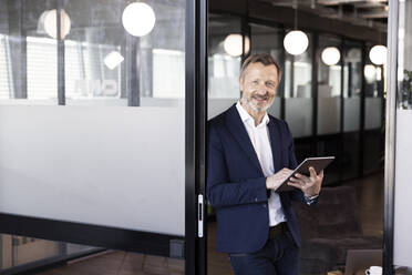 Smiling male professional holding digital tablet while leaning on glass door at office - FKF04164