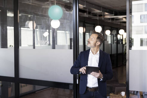 Männlicher Unternehmer mit digitalem Tablet, der an der Tür im Büro steht und nach oben schaut - FKF04166