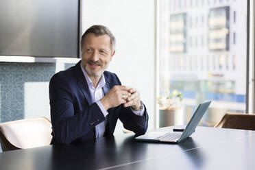 Smiling male entrepreneur looking away while sitting at desk in office - FKF04171