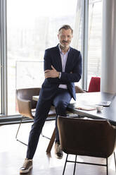 Businessman with arms crossed sitting on desk at office - FKF04177