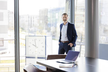 Male professional with hands in pockets standing by clock in office - FKF04183