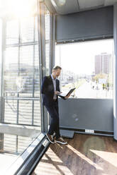 Businessman with laptop leaning on window at office - FKF04193
