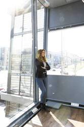 Businesswoman with arms crossed leaning on window at office - FKF04197