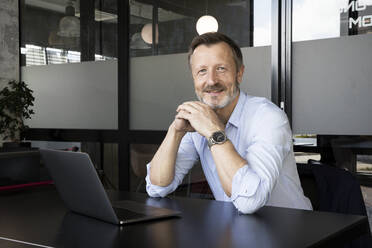 Businessman with hands clasped smiling while sitting at desk in office - FKF04205