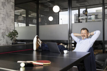 Male professional with hands behind head relaxing at table tennis table in office - FKF04206