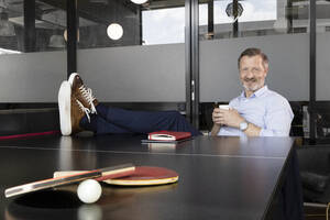 Lächelnder Geschäftsmann, der Kaffee trinkt, während er an der Tischtennisplatte im Büro sitzt - FKF04207