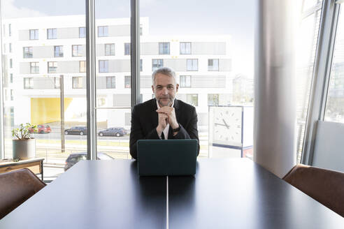 Männlicher Unternehmer mit verschränkten Händen vor einem Laptop im Büro sitzend - FKF04211