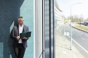 Männlicher Unternehmer schaut weg und hält einen Laptop vor einer Wand im Büro - FKF04216