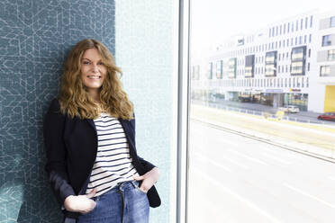 Weibliche Unternehmerin lächelt, während sie sich im Büro an die Wand am Fenster lehnt - FKF04227