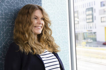 Happy businesswoman with brown hair leaning on wall at office - FKF04228