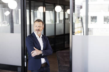 Male professional with arms crossed looking away while standing in front of glass door at office - FKF04271