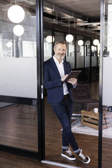 Lächelnder männlicher Unternehmer, der einen Laptop hält und sich an eine Glastür im Büro lehnt - FKF04274