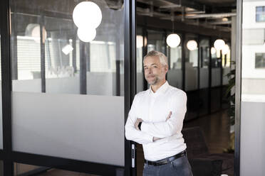 Mature male professional with arms crossed leaning on glass door at office - FKF04275