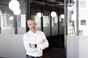 Mature male entrepreneur with arms crossed leaning on glass door at office - FKF04277