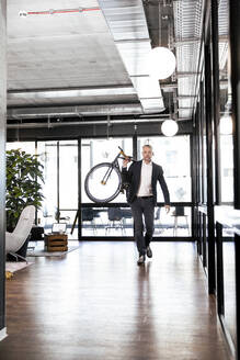 Geschäftsmann trägt Fahrrad auf der Schulter, während er im Büro spazieren geht - FKF04287