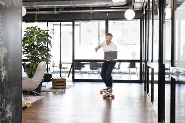 Smiling male professional with laptop riding skateboard at office - FKF04289