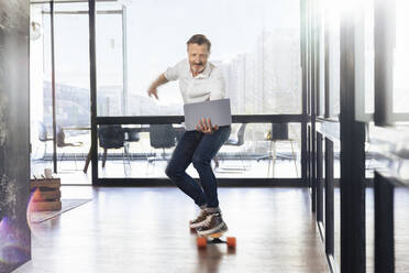 Älterer Geschäftsmann auf dem Skateboard mit Blick auf den Laptop im Büro - FKF04290