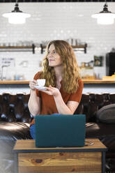 Geschäftsfrau trinkt Kaffee, während sie in der Büro-Cafeteria vor ihrem Laptop sitzt - FKF04303