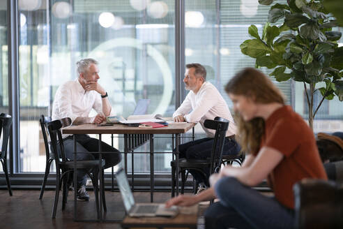 Männliche Unternehmer diskutieren im Hintergrund, während eine Geschäftsfrau im Büro am Laptop arbeitet - FKF04332