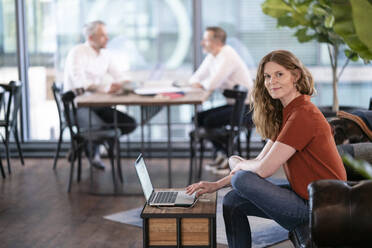 Geschäftsfrau vor einem Laptop sitzend mit männlichen Kollegen, die im Hintergrund in einer Büro-Cafeteria diskutieren - FKF04333