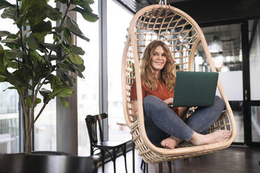 Smiling businesswoman with laptop sitting on swing chair at office cafeteria - FKF04337