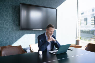 Businessman with hands clasped sitting in front of laptop at table in office - FKF04351